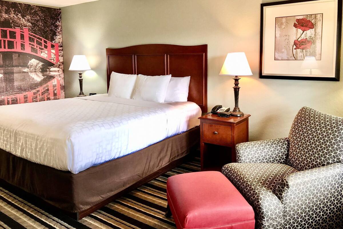A hotel room with a large bed, two bedside tables with lamps, a patterned armchair, and a framed picture on the wall.