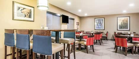 Un comedor de hotel con varias mesas y sillas, un televisor montado en la pared y obras de arte enmarcadas en las paredes.