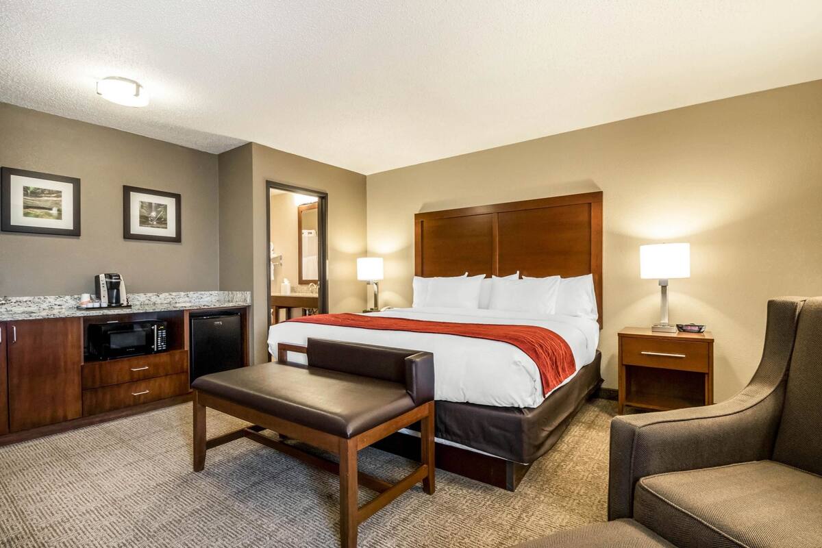 A hotel room with a bed, a desk with a microwave, a chair, a small table, and a nightstand with a lamp.