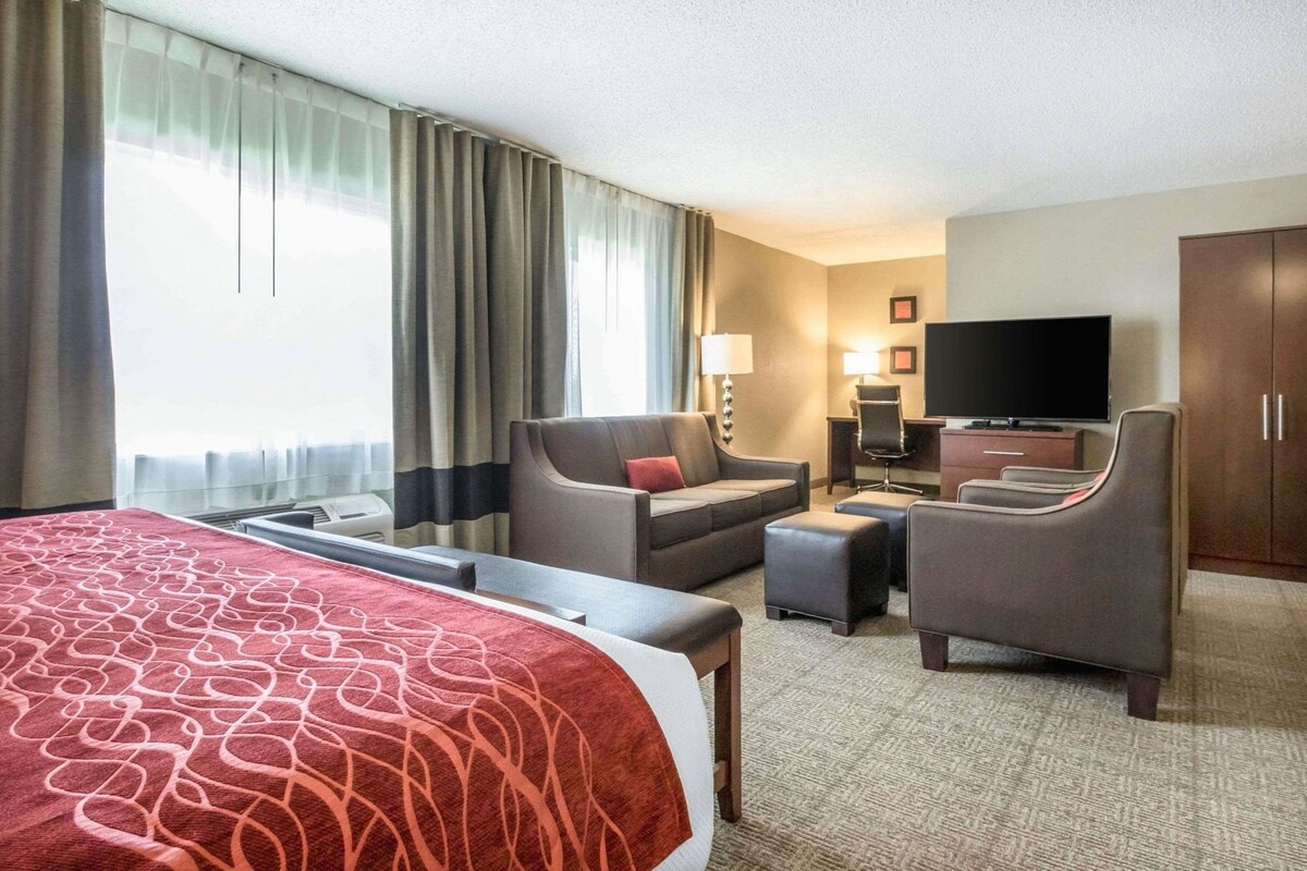 A hotel room with a large bed, two armchairs, a desk with a TV, and a window with curtains.