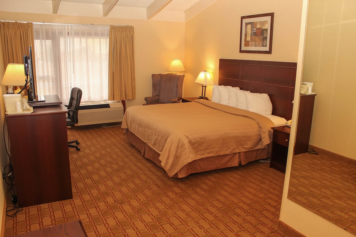 A hotel room with a large bed, two bedside lamps, a desk with a computer, a chair, and a window with curtains.