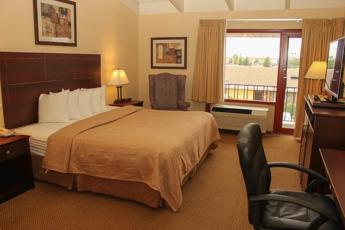 A hotel room with a large bed, a desk with a computer, a chair, a window with curtains, and a painting on the wall.