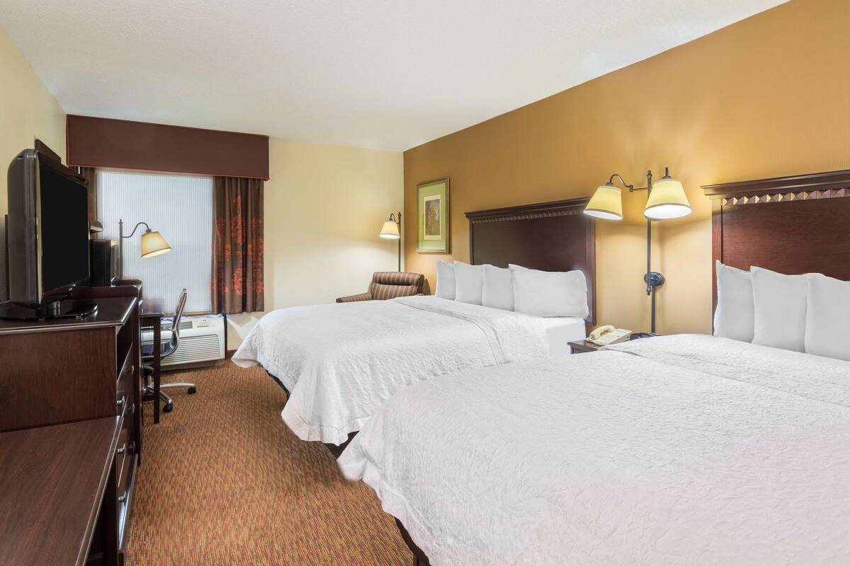 A hotel room with two beds, a desk with a computer, a chair, a lamp, and a framed picture on the wall.