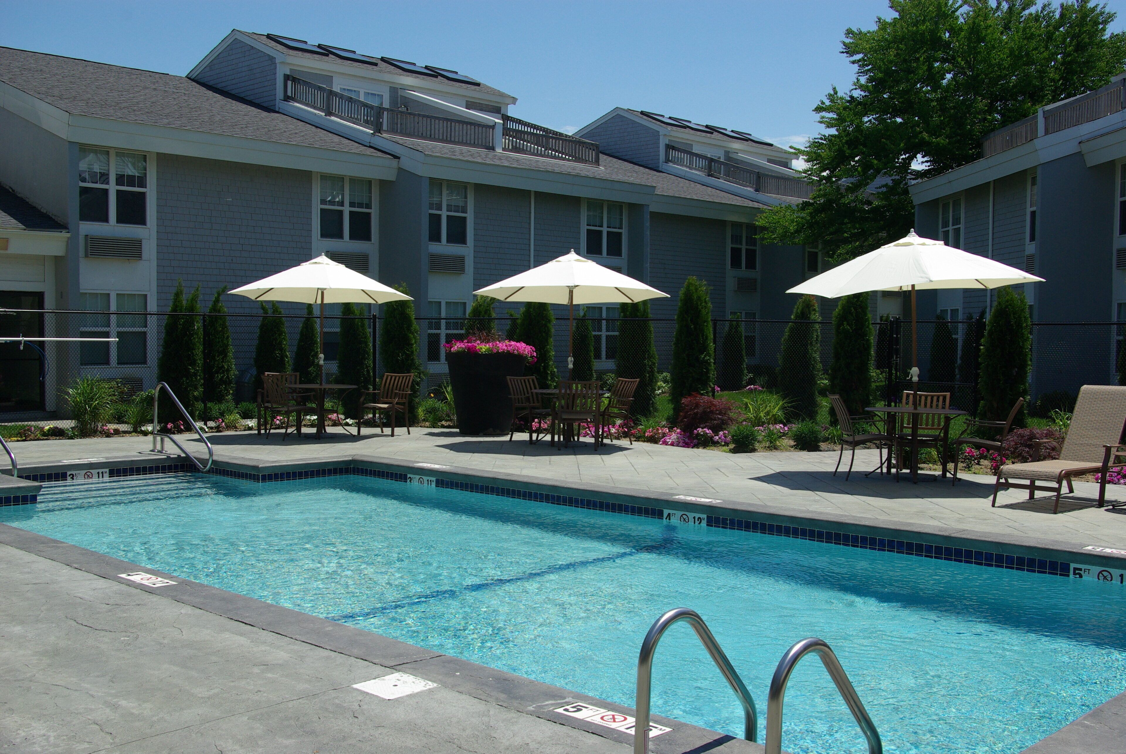 A swimming pool with a metal ladder, surrounded by lounge chairs and umbrellas, with a building in the background.