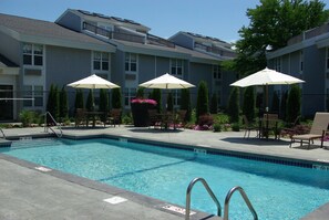 A swimming pool with a metal ladder, surrounded by lounge chairs and umbrellas, with a building in the background.