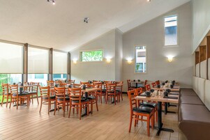 A spacious dining area with multiple tables and chairs, large windows, and wall-mounted lighting.