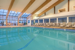 An indoor swimming pool with clear blue water, wooden beams on the ceiling, and lounge chairs along the poolside.