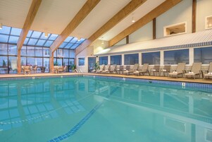 An indoor swimming pool with clear blue water, wooden beams on the ceiling, and lounge chairs along the poolside.