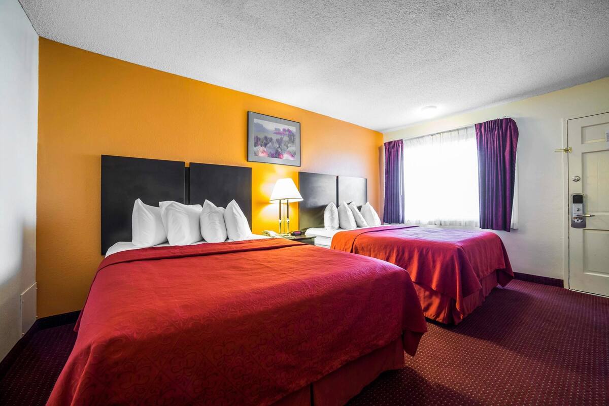 A hotel room with two beds, a nightstand with a lamp, a painting on the wall, and a window with curtains.