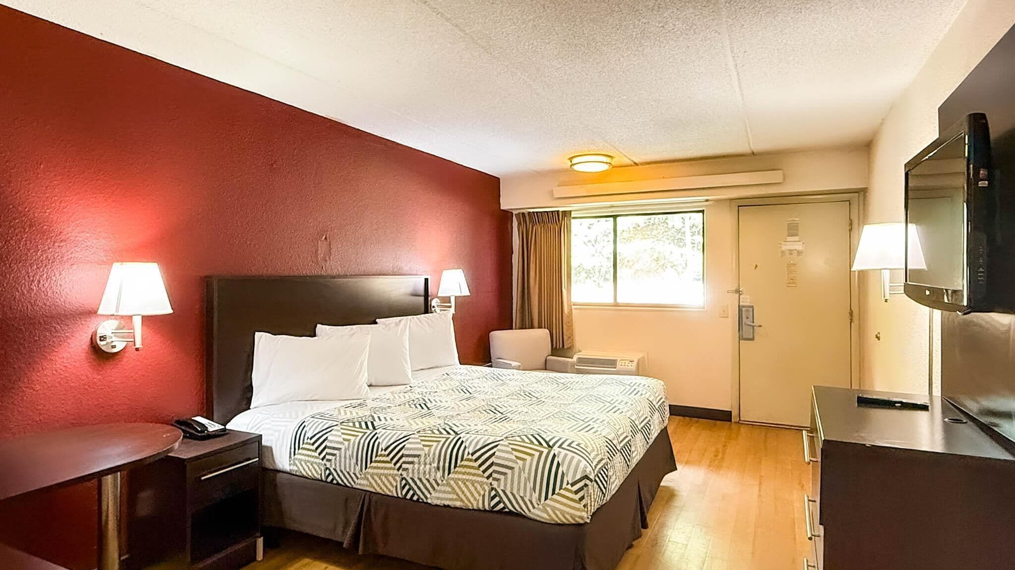 Habitación de hotel con cama, lámparas de noche, escritorio, silla, televisor y ventana con cortinas.