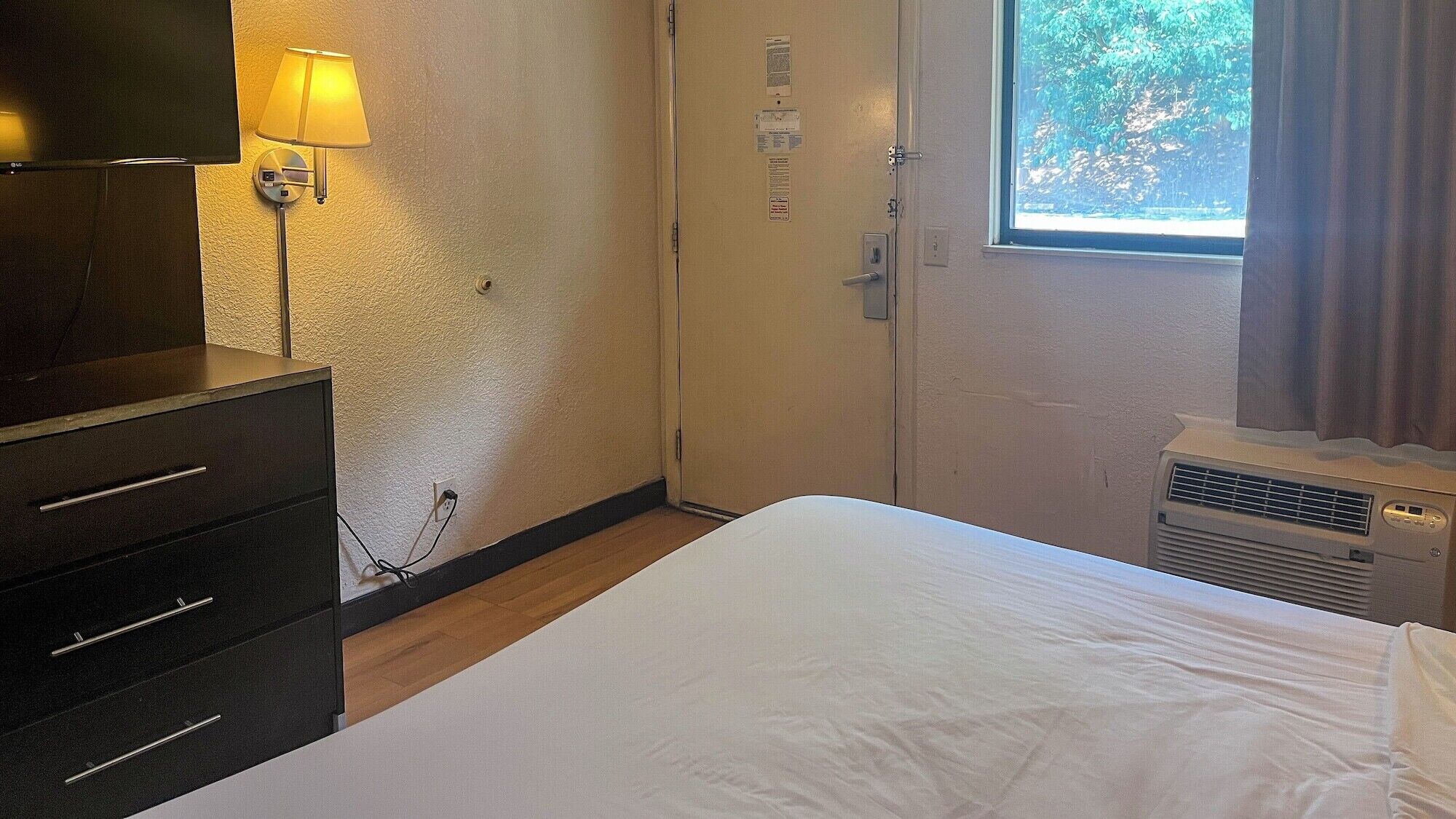 A hotel room with a bed, a dresser, a lamp, a window with curtains, and a wall-mounted heater.