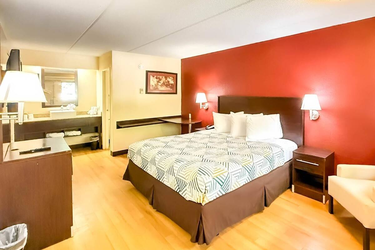 A hotel room with a bed, bedside tables, a chair, a desk, a mirror, and a red accent wall.