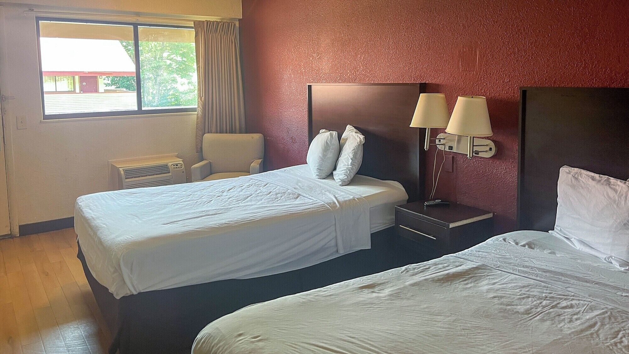 A hotel room with a large bed, a smaller bed, a chair, a nightstand with lamps, and a window with curtains.