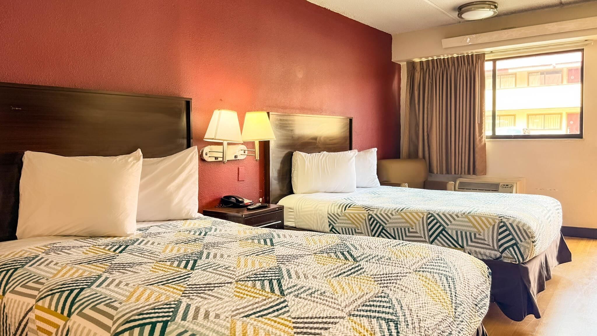Habitación de hotel con dos camas, una mesita de noche, una lámpara y un ventanal con cortinas.