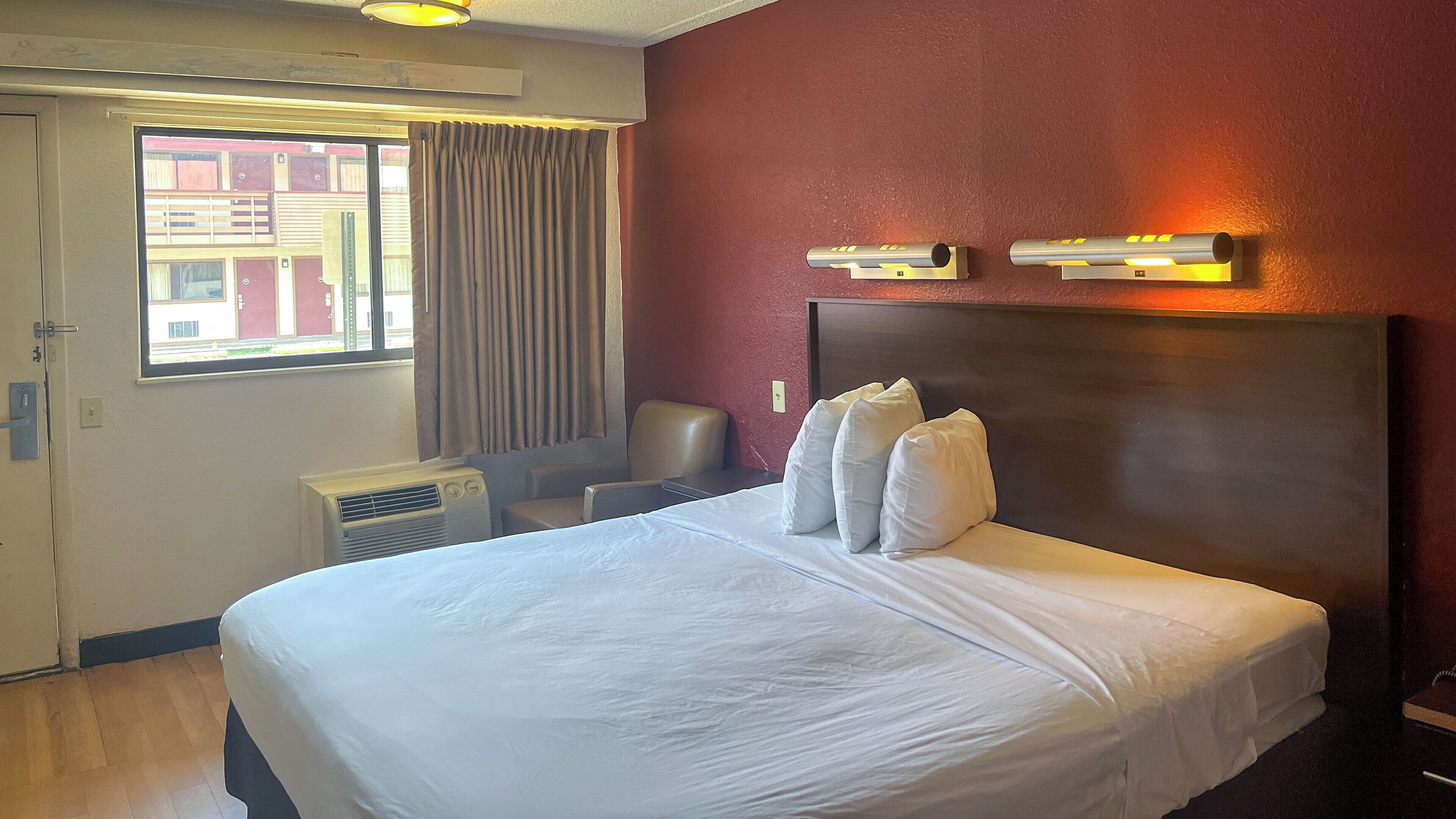 A hotel room with a large bed, an air conditioning unit, a window with curtains, and a wall-mounted light fixture.