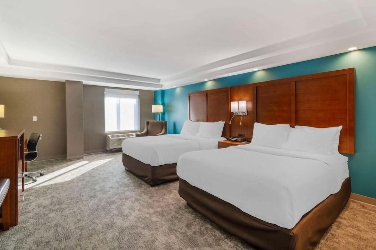 A hotel room with two beds, a desk, a chair, a window, and a wall-mounted lamp.