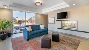A modern hotel lobby with a blue sofa, large windows, a flat-screen TV, and a patterned rug.