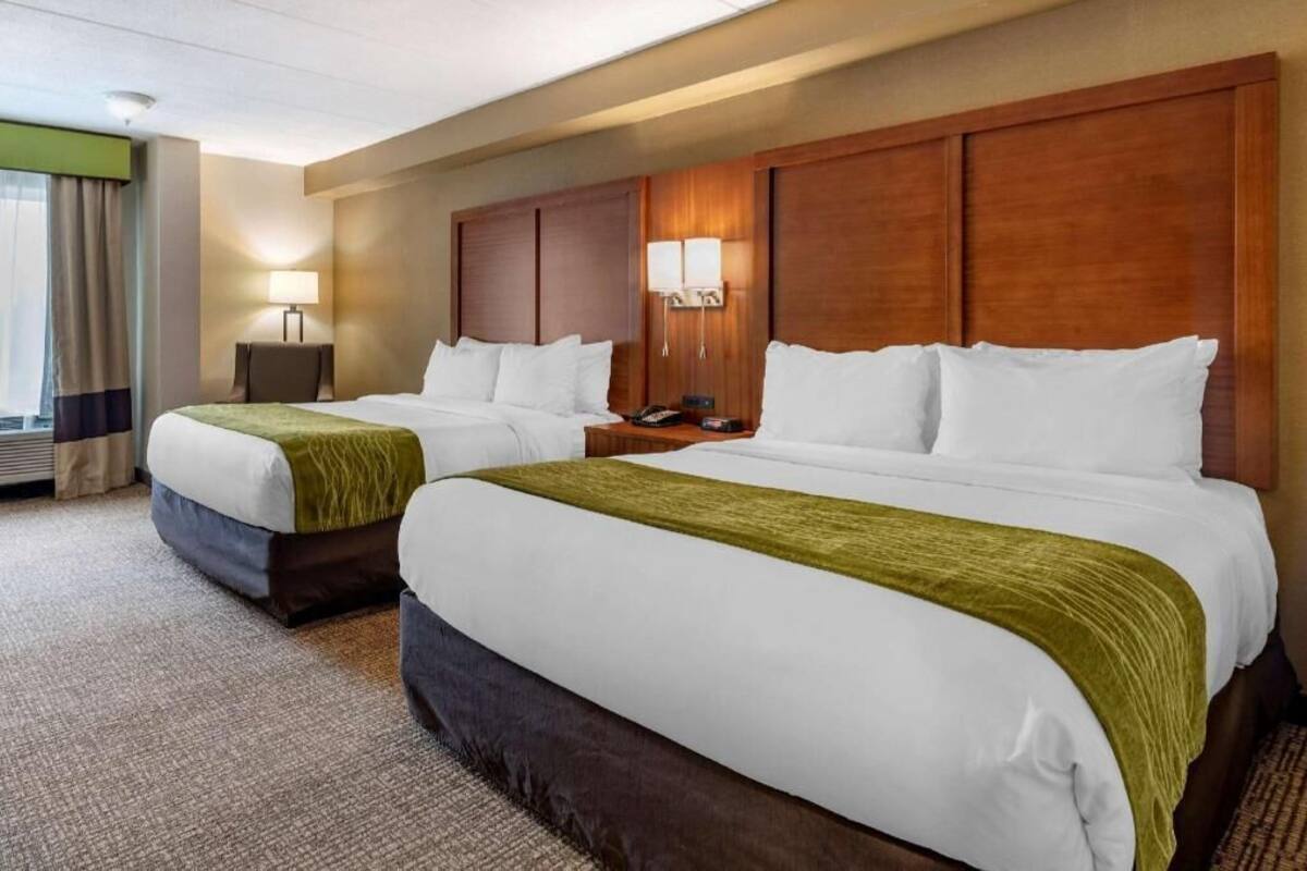 A hotel room with two beds, a wooden headboard, bedside lamps, a desk, and a view of the outdoors.
