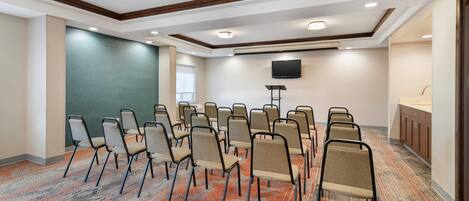 A conference room with rows of chairs, a flat-screen TV, and a kitchenette area.