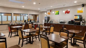 Un restaurante con varias mesas y sillas, una barra con dispensadores de comida y bebida, y vistas a través de amplios ventanales.