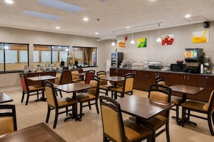 Um restaurante com várias mesas e cadeiras, um balcão com dispensadores de comida e bebida, e vista através de amplas janelas.