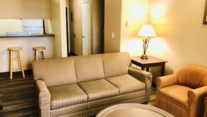 A living room with a striped sofa, a round coffee table, and an orange armchair. There is a small table with a lamp and two wooden stools. A kitchen area is visible in the background.