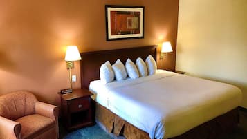 Ein Hotelzimmer mit einem großen Bett, zwei Nachttischlampen, einem gerahmten Kunstwerk über dem Bett, einem kleinen Nachttisch, einem braunen Sessel und einem Teppichboden.