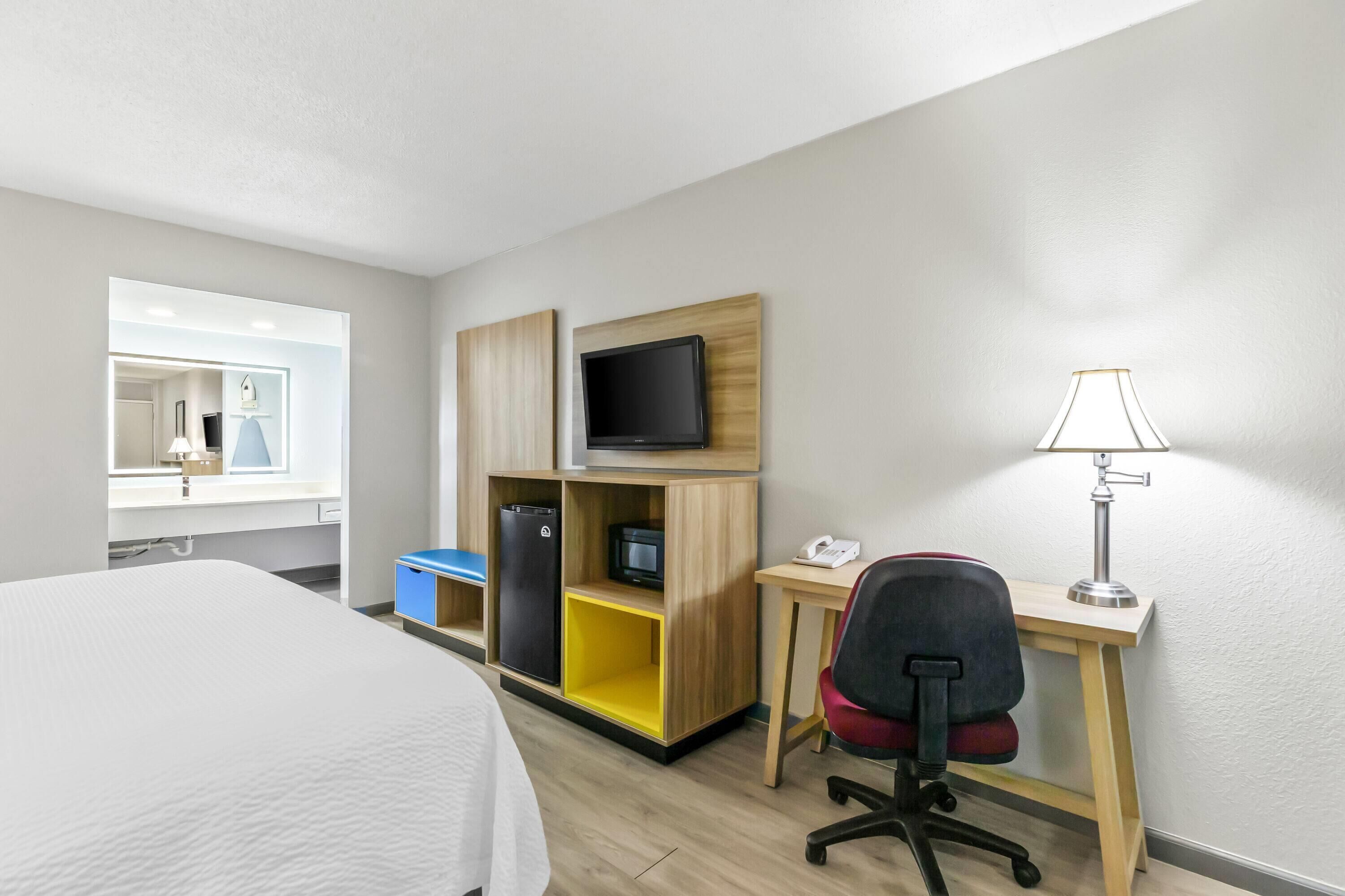 A hotel room with a bed, a desk with a lamp, a TV, a refrigerator, and a chair.