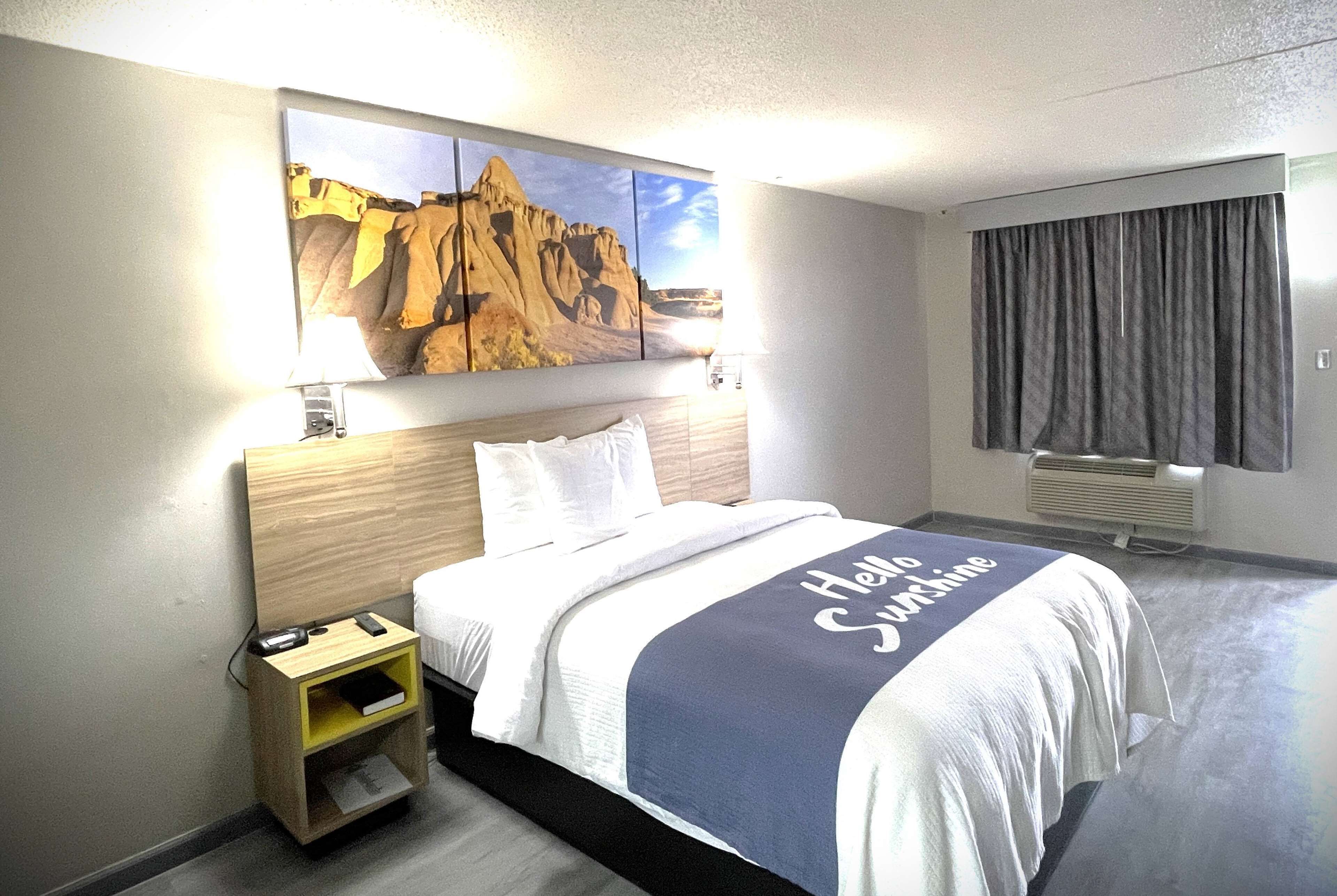 A hotel room with a bed, a nightstand, a painting on the wall, a window with curtains, and a view of a mountain.