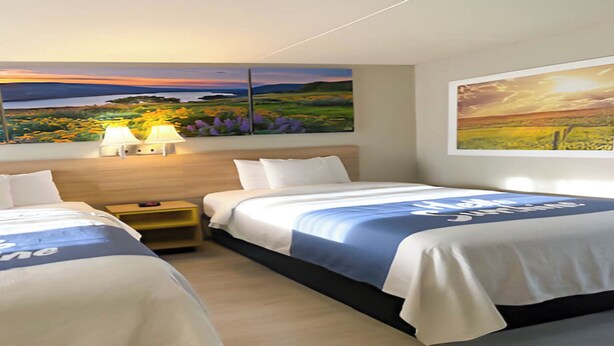 A hotel room with two beds, a nightstand, a lamp, and two large landscape paintings on the walls.