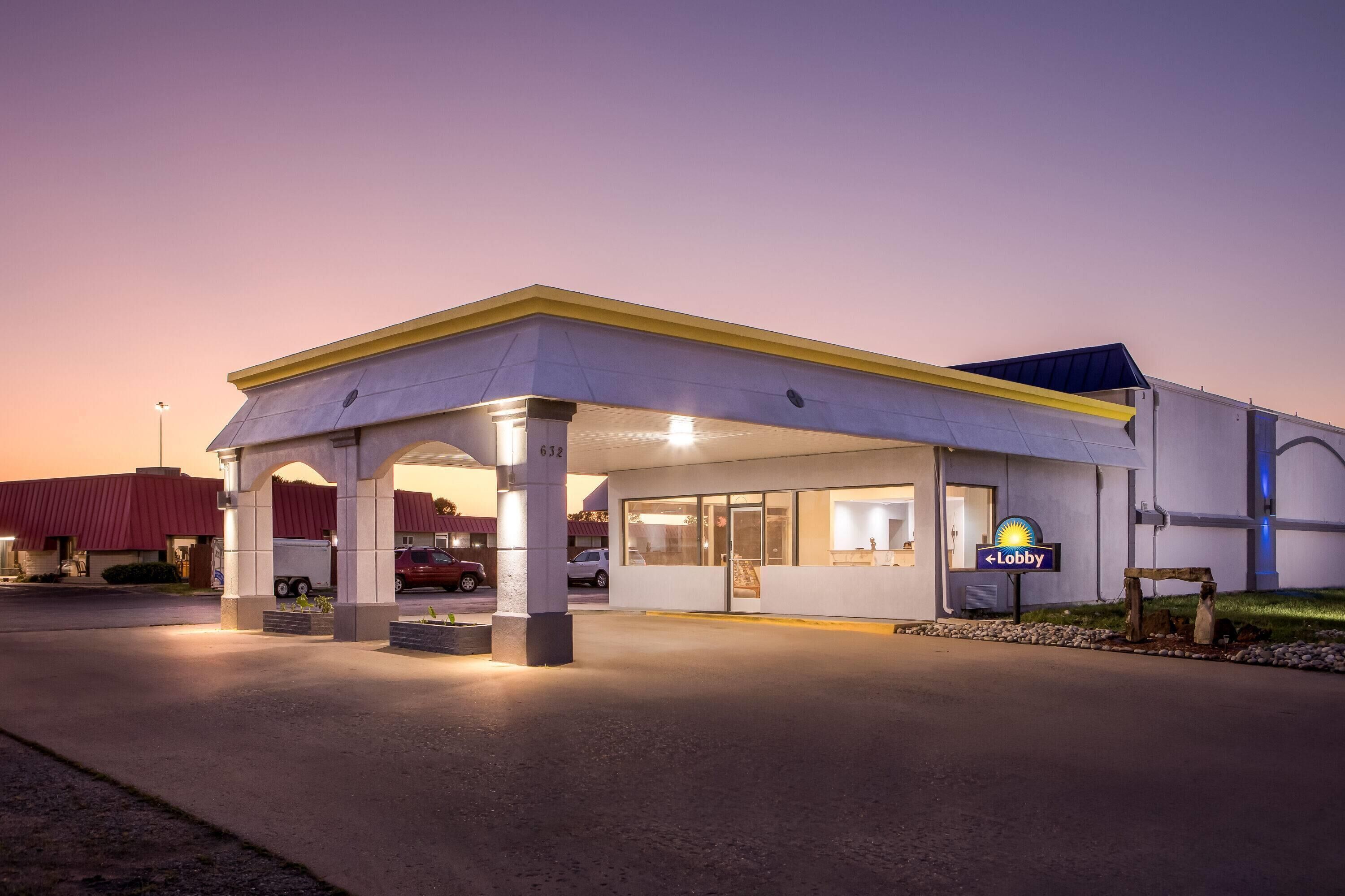 Een hotel met een overdekte ingang, een zichtbaar bord dat een receptie aangeeft, en een parkeerplaats met enkele auto's.