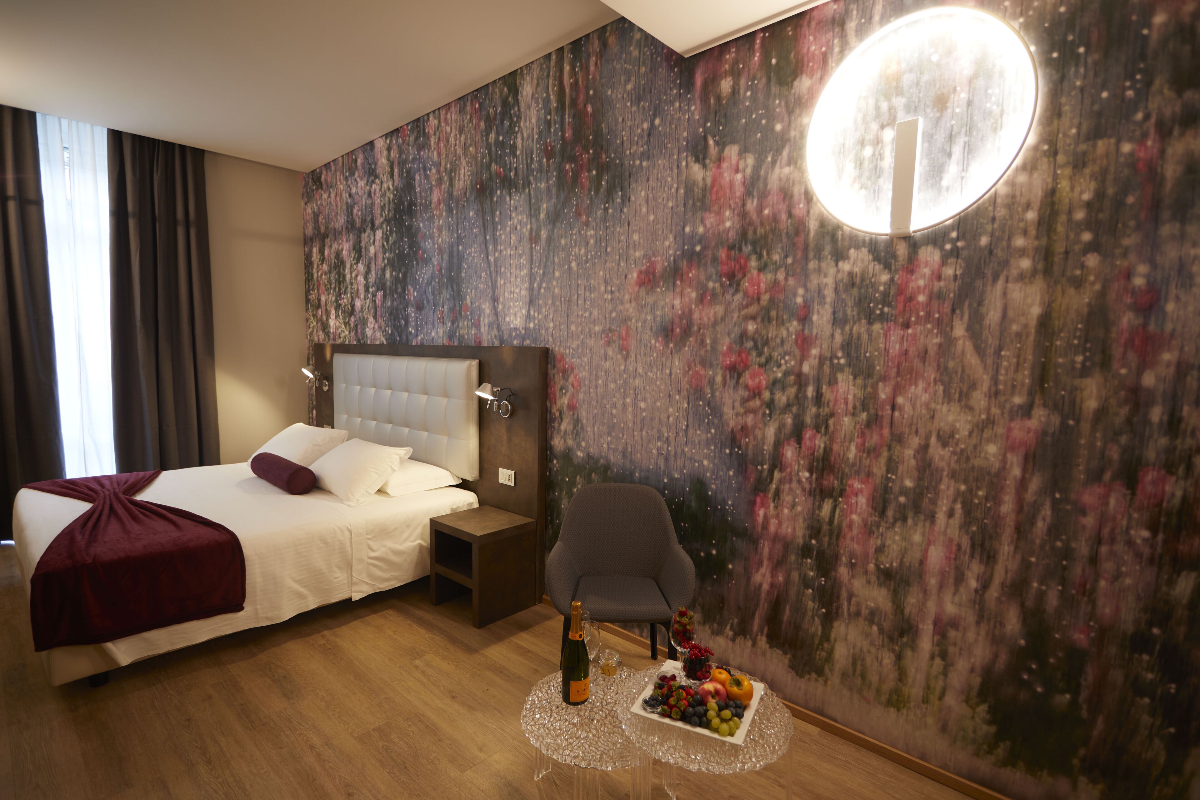 Habitación de hotel con una cama grande, una mesita de noche, una silla, un mural en la pared y una pequeña mesa con una botella y fruta.