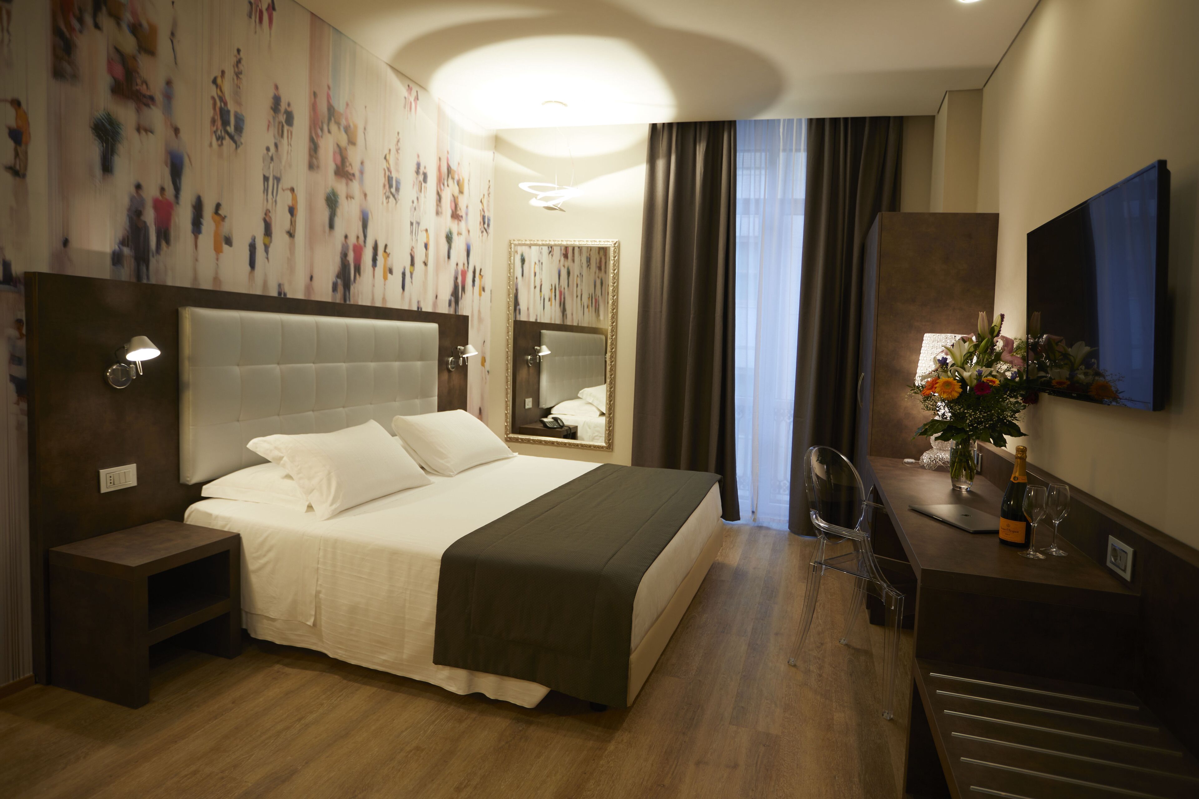 Ein Hotelzimmer mit einem großen Bett, einem Nachttisch, einem Schreibtisch mit Stuhl, einem Spiegel und einem an der Wand befestigten Fernseher.