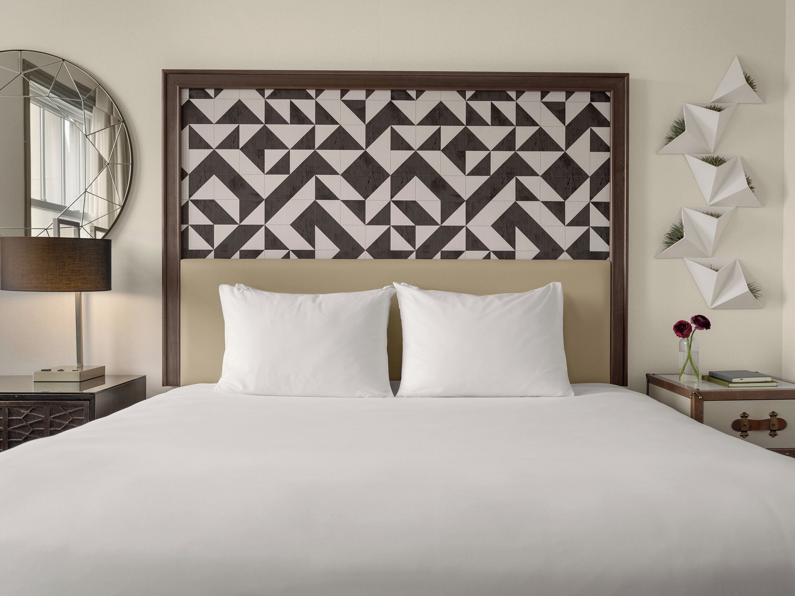 A neatly made bed with white bedding, a geometric patterned headboard, a bedside lamp, and a small table with a vase of flowers.