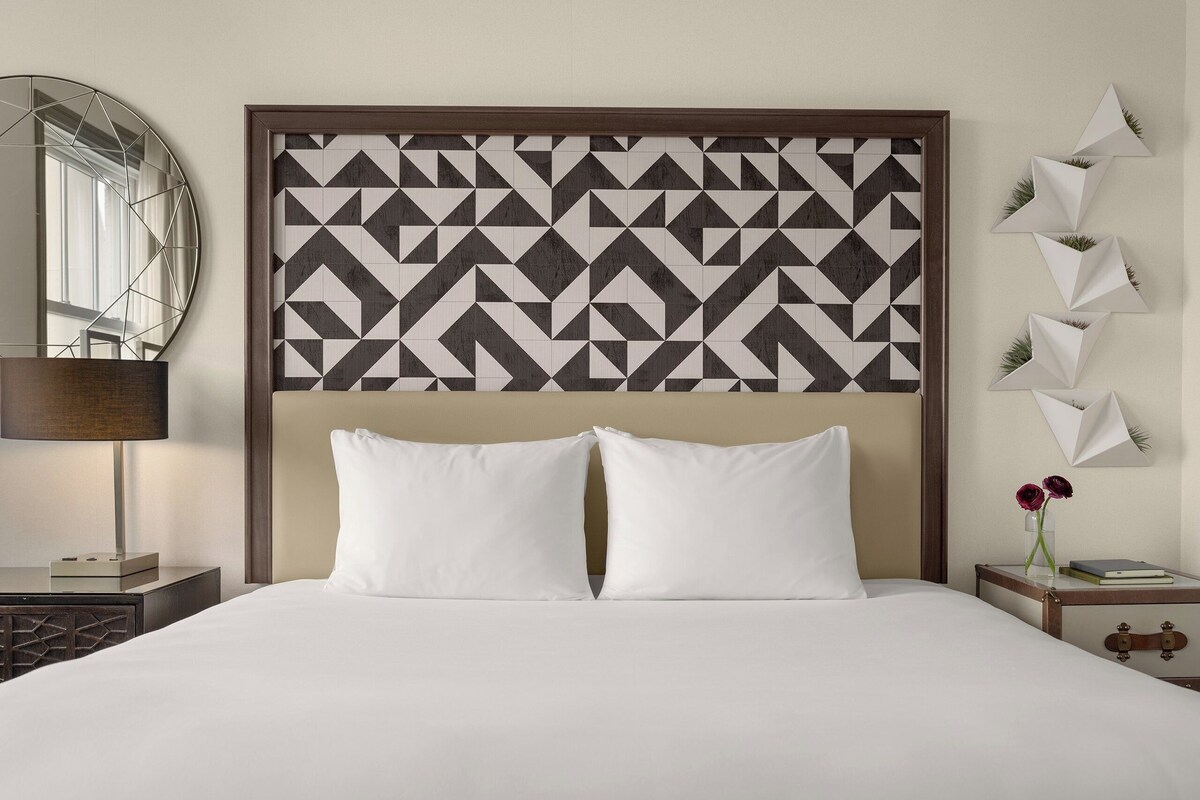 A neatly made bed with white bedding, a geometric patterned headboard, a bedside lamp, and a small table with a vase of flowers.