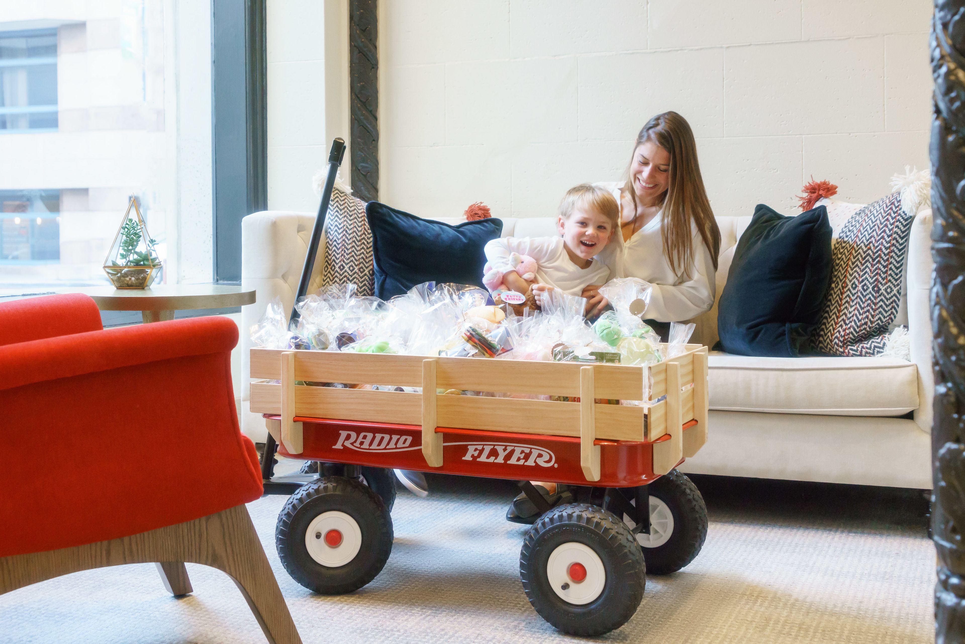 A room with a wooden toy wagon labeled "Radio Flyer" containing various items, a red chair, and a white sofa with patterned cushions.