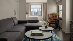 A modern living room with a grey sofa, a round coffee table with books, a brown leather armchair, and a view of the city through large windows.