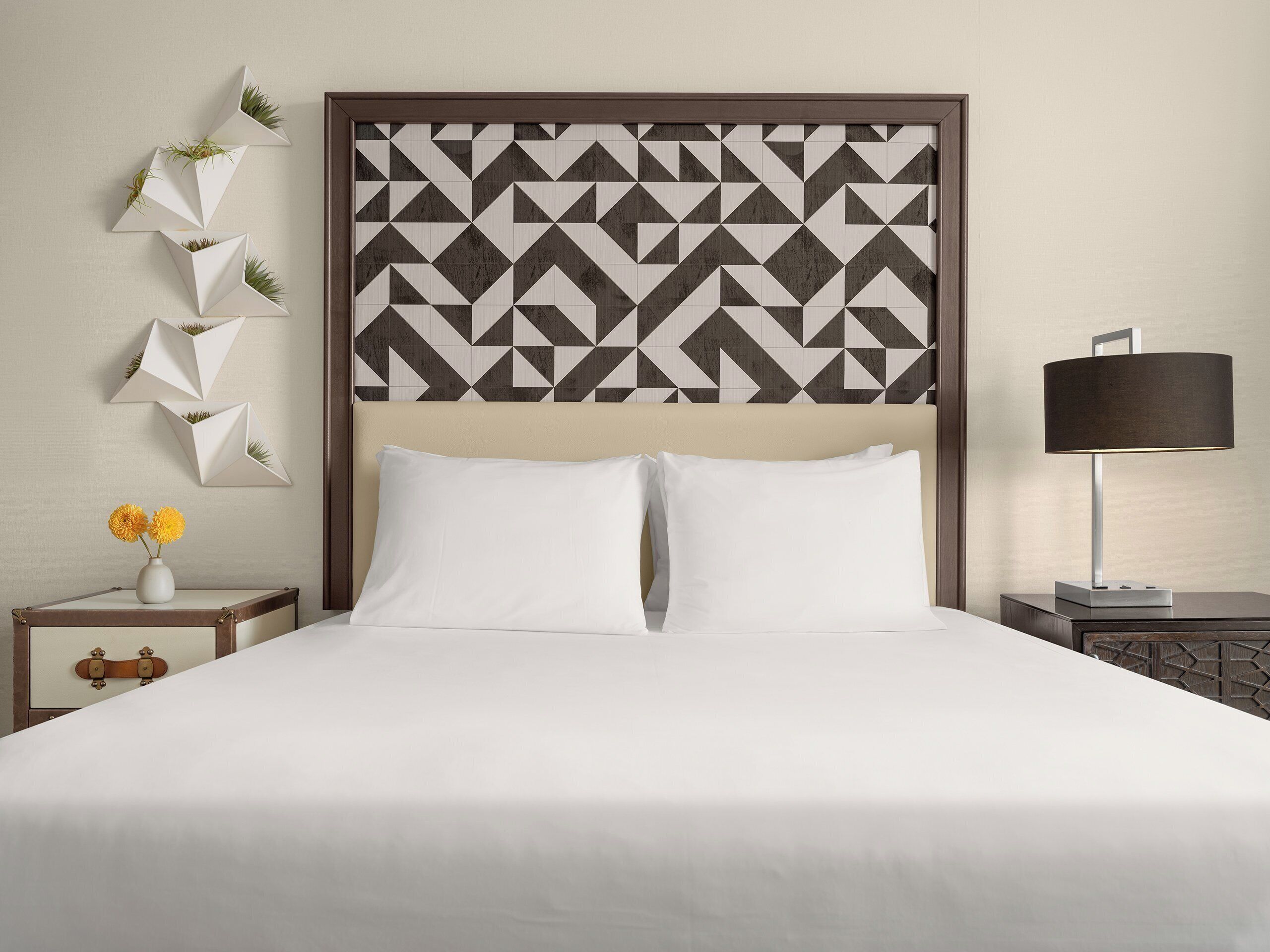 A hotel room with a bed, a nightstand with a lamp, and a wall with geometric patterned wallpaper.