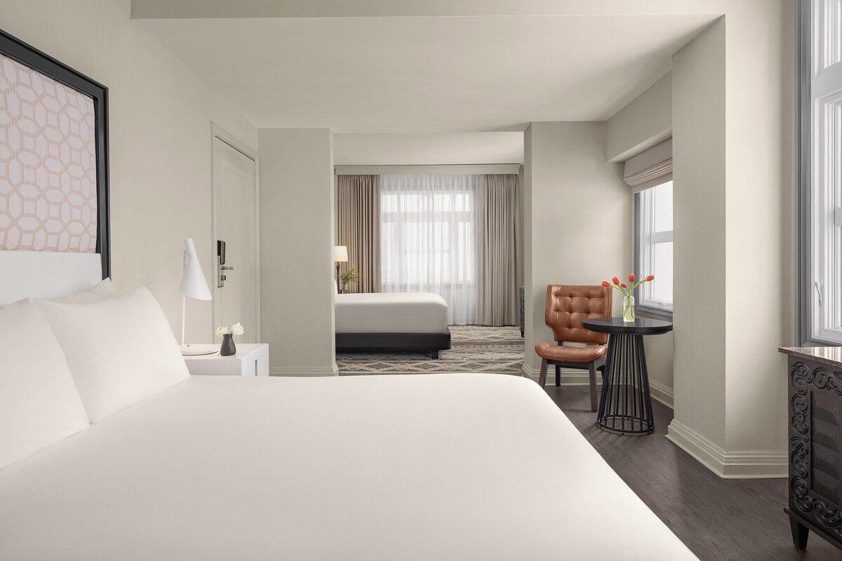 Hotellihuone, jossa on suuri sänky, työpöytä tuolilla, ikkuna verhoilla ja näkymä käytävälle.