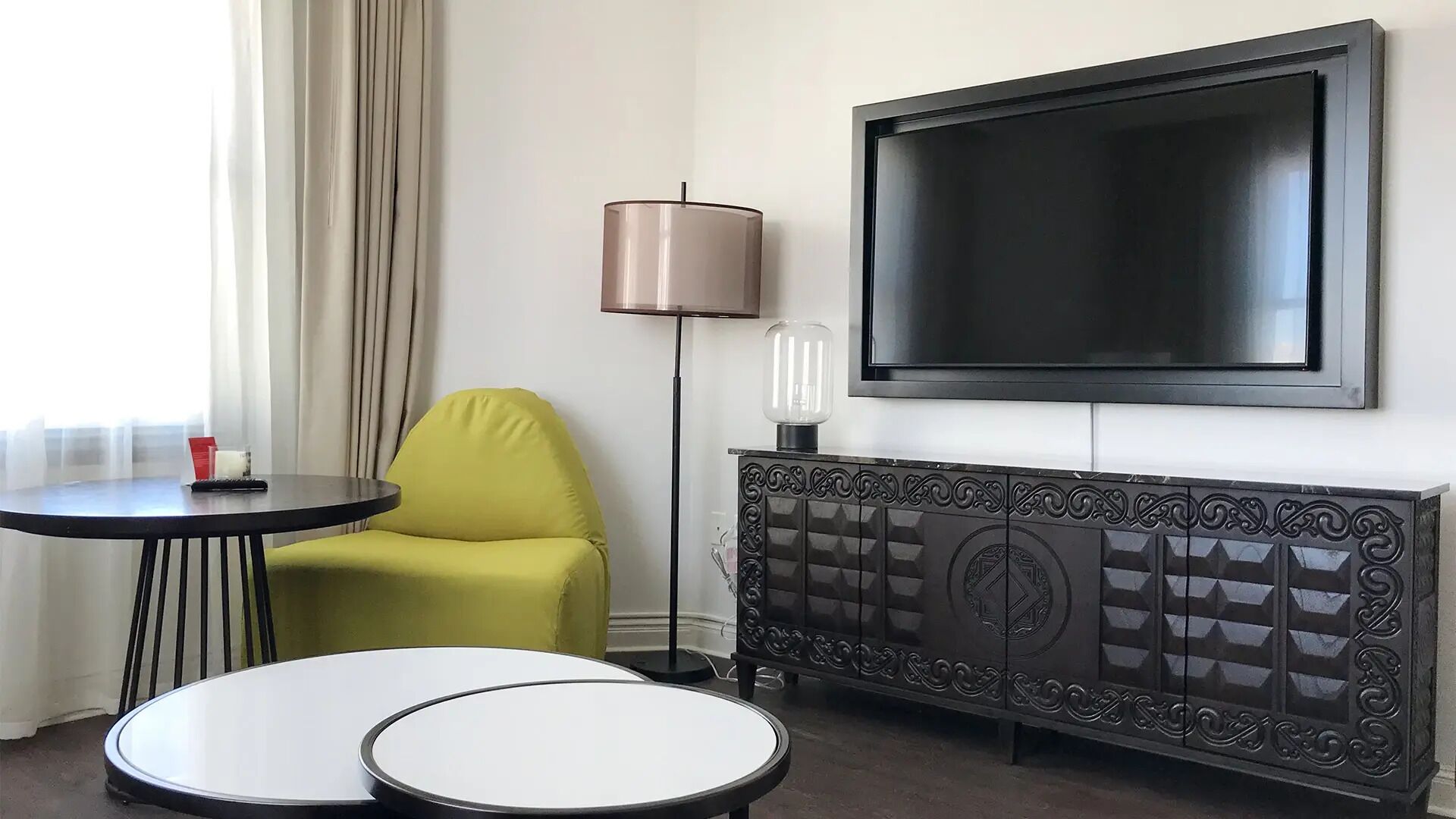 A hotel room with a TV mounted on the wall, a lamp, a sofa, and two small tables.