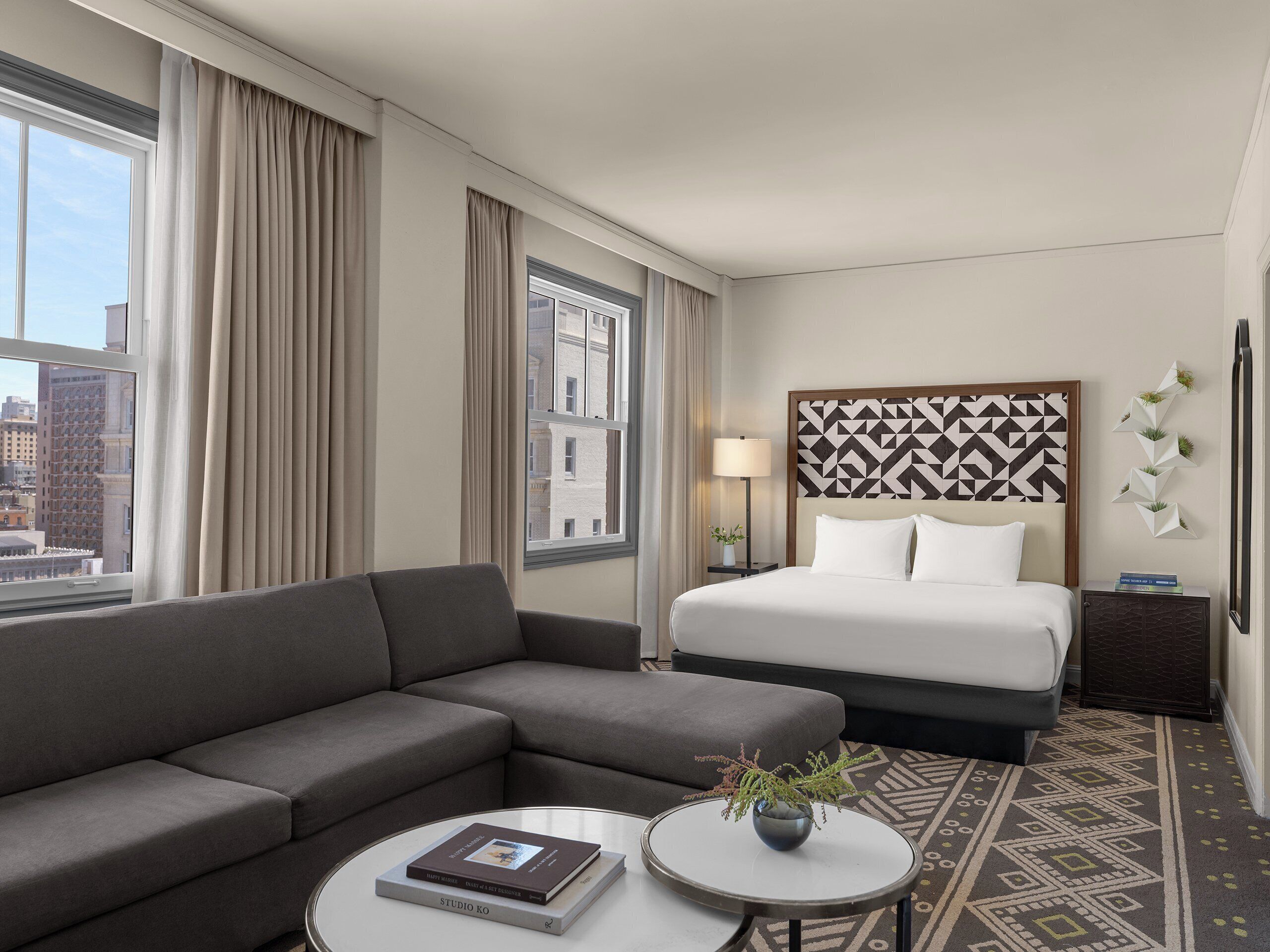 Een moderne hotelkamer met een grijze bank, een ronde salontafel met een boek en een vaas, een bed met witte lakens, een gestreept hoofdeinde en uitzicht op de stad door grote ramen.