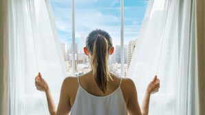 A person standing by a window with sheer curtains, looking out at a cityscape.