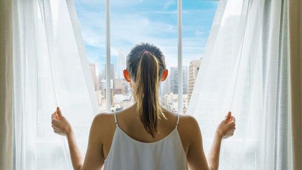 A person standing by a window with sheer curtains, looking out at a cityscape. - Hotel Spero, Vignette Collection by IHG (San Francisco)