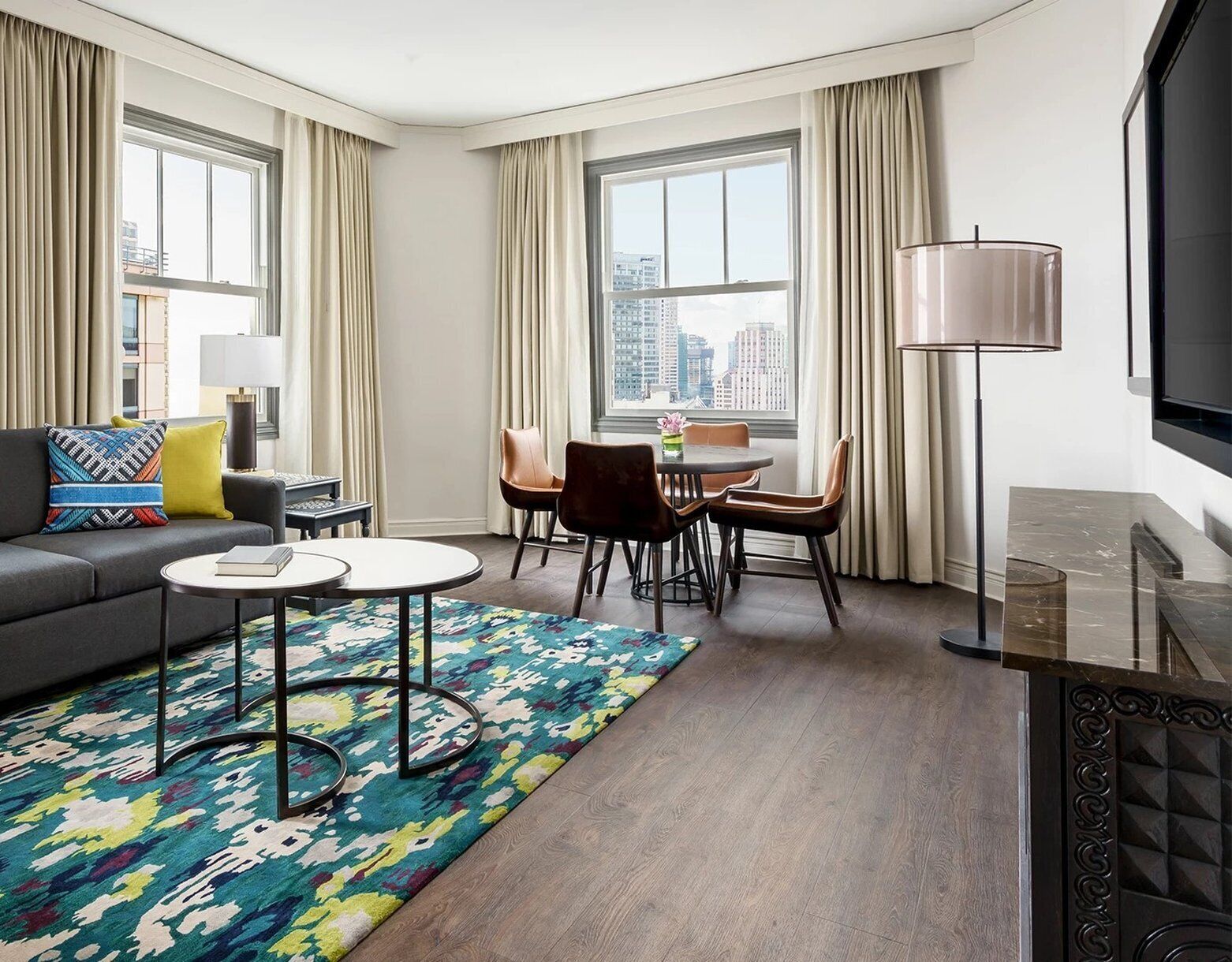 A modern living room with a sofa, coffee table, and dining area. There are large windows with curtains, a city view, and a flat-screen TV.