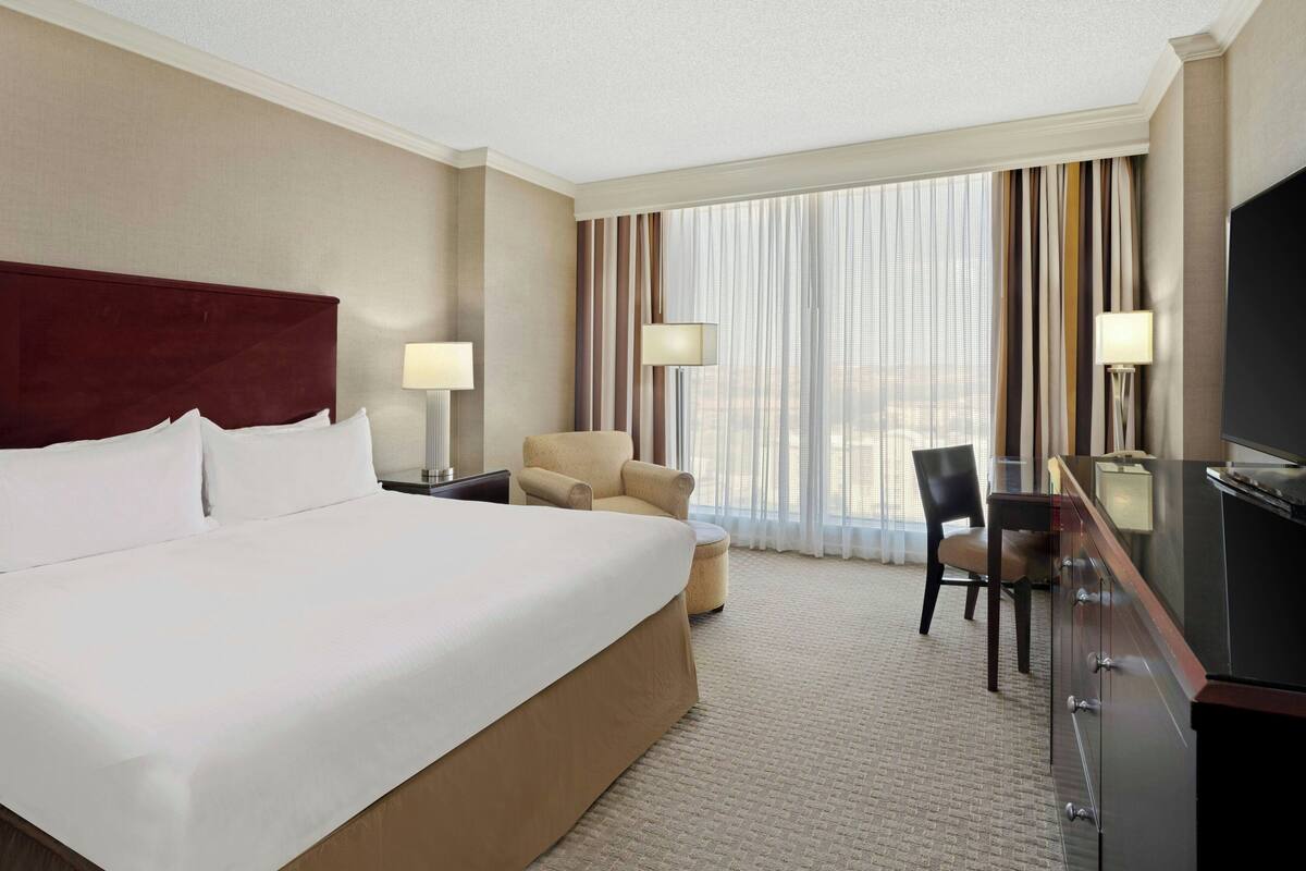 Habitación de hotel con una cama grande, un escritorio, dos sillas, un televisor y una ventana con cortinas.