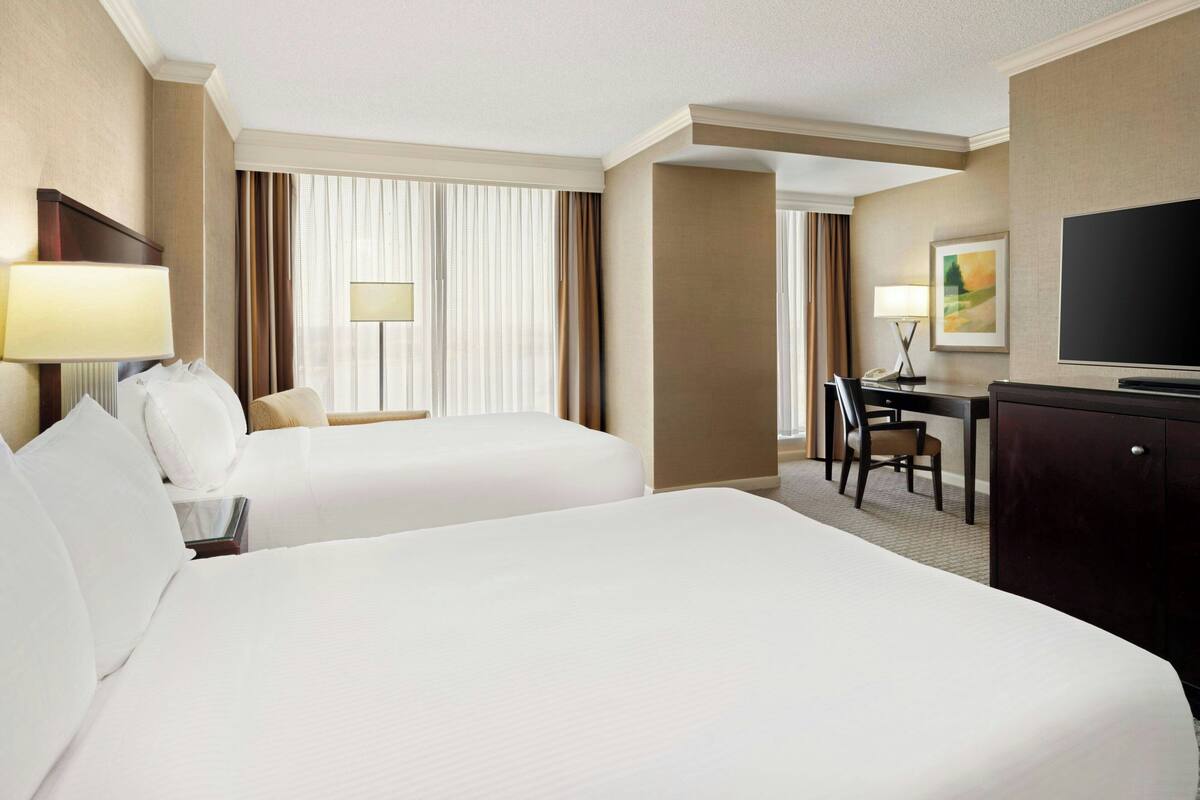 Habitación de hotel con dos camas, un escritorio, un televisor y un ventanal con cortinas.
