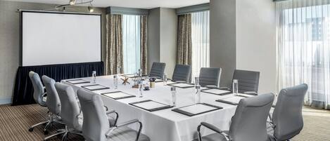 Un salón de conferencias con una larga mesa, seis sillas, una pantalla de proyector y un amplio ventanal con cortinas.