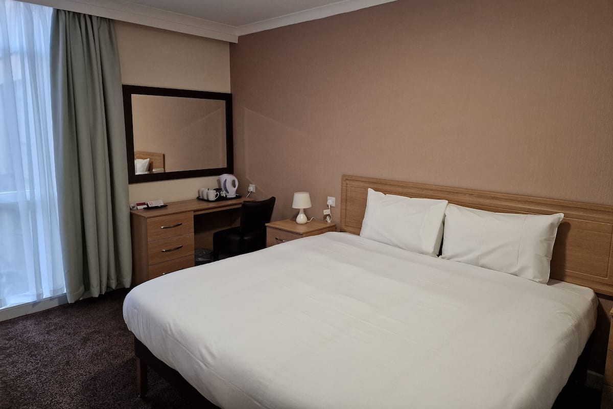 Habitación de hotel con una cama grande, un escritorio con silla, un espejo y una mesita de noche con lámpara.