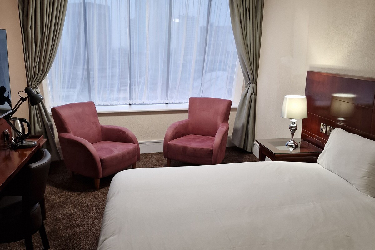 Een hotelkamer met een groot bed, twee fauteuils, een bureau met lamp en een nachtkastje met lamp.