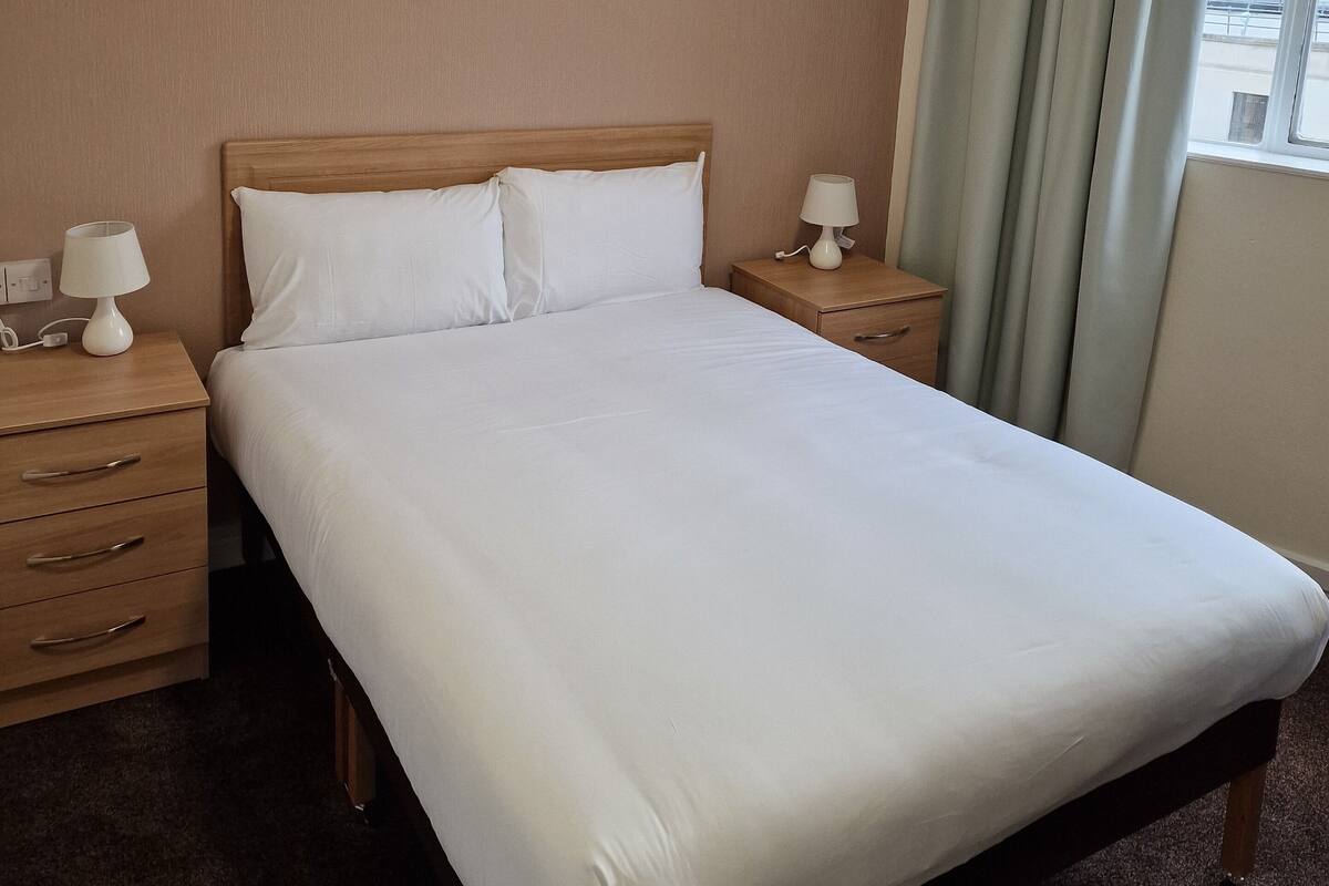 Habitación de hotel con una cama grande, dos mesitas de noche con lámparas, un cabecero de madera y vistas a la ciudad desde la ventana.
